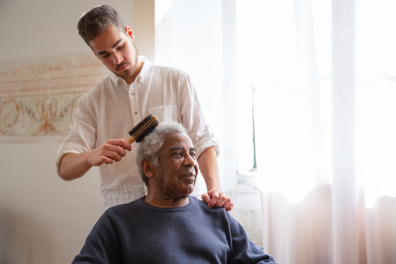 Young man gently combs elderly man's hair by a sunlit window, portraying caregiving and family bond.